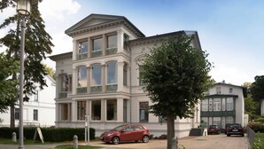 A large, white, multi-story building with a classical architectural style, featuring large windows and a balcony.