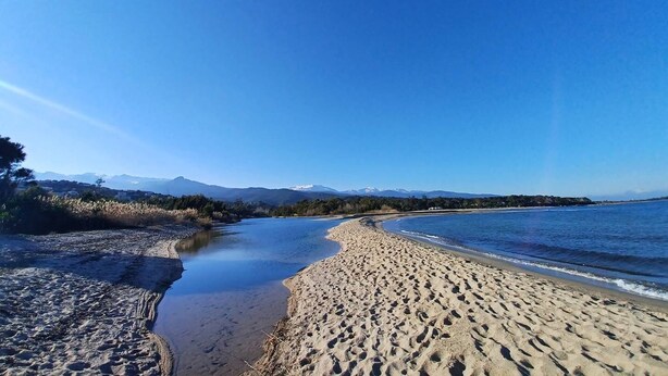 Ein Strand mit blauem Himmel, einem Fluss, der ins Meer fließt, und Bergen im Hintergrund.