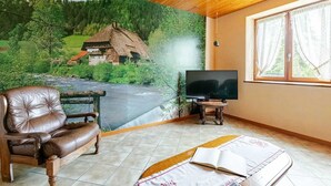 A room with a mural of a rural landscape, a leather sofa, a television on a wooden stand, and a bed with a book.