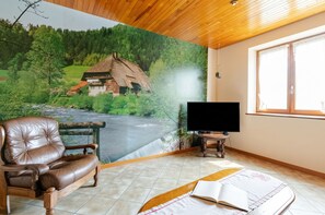 A room with a mural of a rural landscape, a leather armchair, a television on a stand, and a window with curtains. - Homerez - Apartment in Epfig (Epfig)