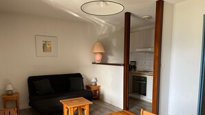 Un espace de vie compact comprenant un canapé noir, une table à manger en bois et un coin cuisine visible à travers une porte ouverte.