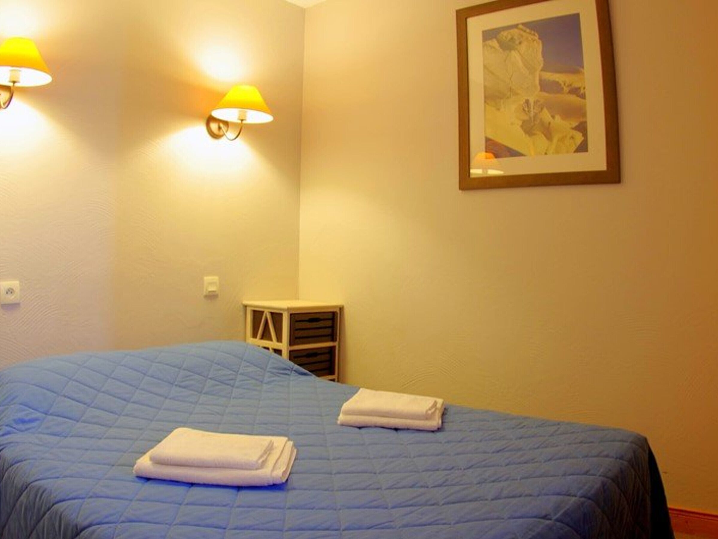 Une chambre d’hôtel avec une couvre-lit bleue, deux serviettes blanches, un tableau encadré au mur et deux lampes fixées au mur.