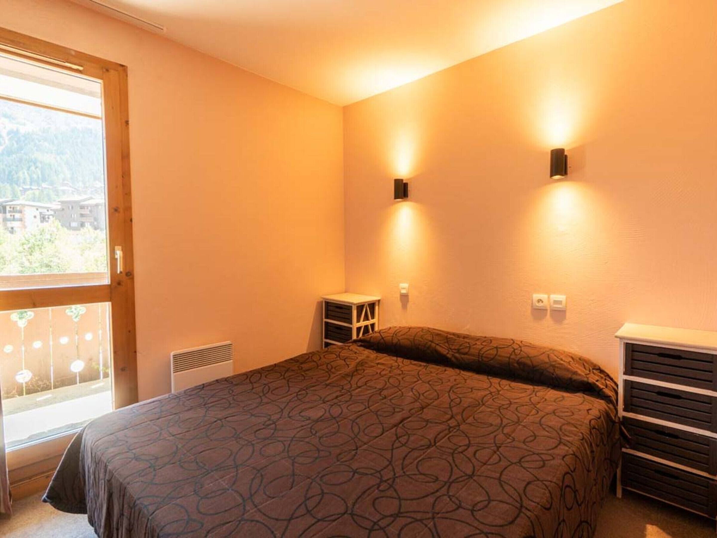 A bedroom with a bed, a small bedside table, a heater, and a window with a view of buildings.