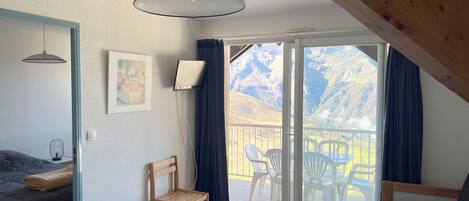 A room with a bed, a wooden chair, a small table, and a balcony with a view of mountains.
