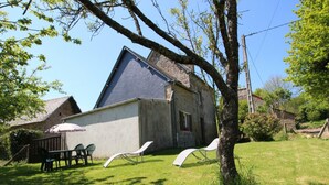 Un bâtiment en pierre avec un toit en pente, une cour verdoyante et deux chaises longues blanches.