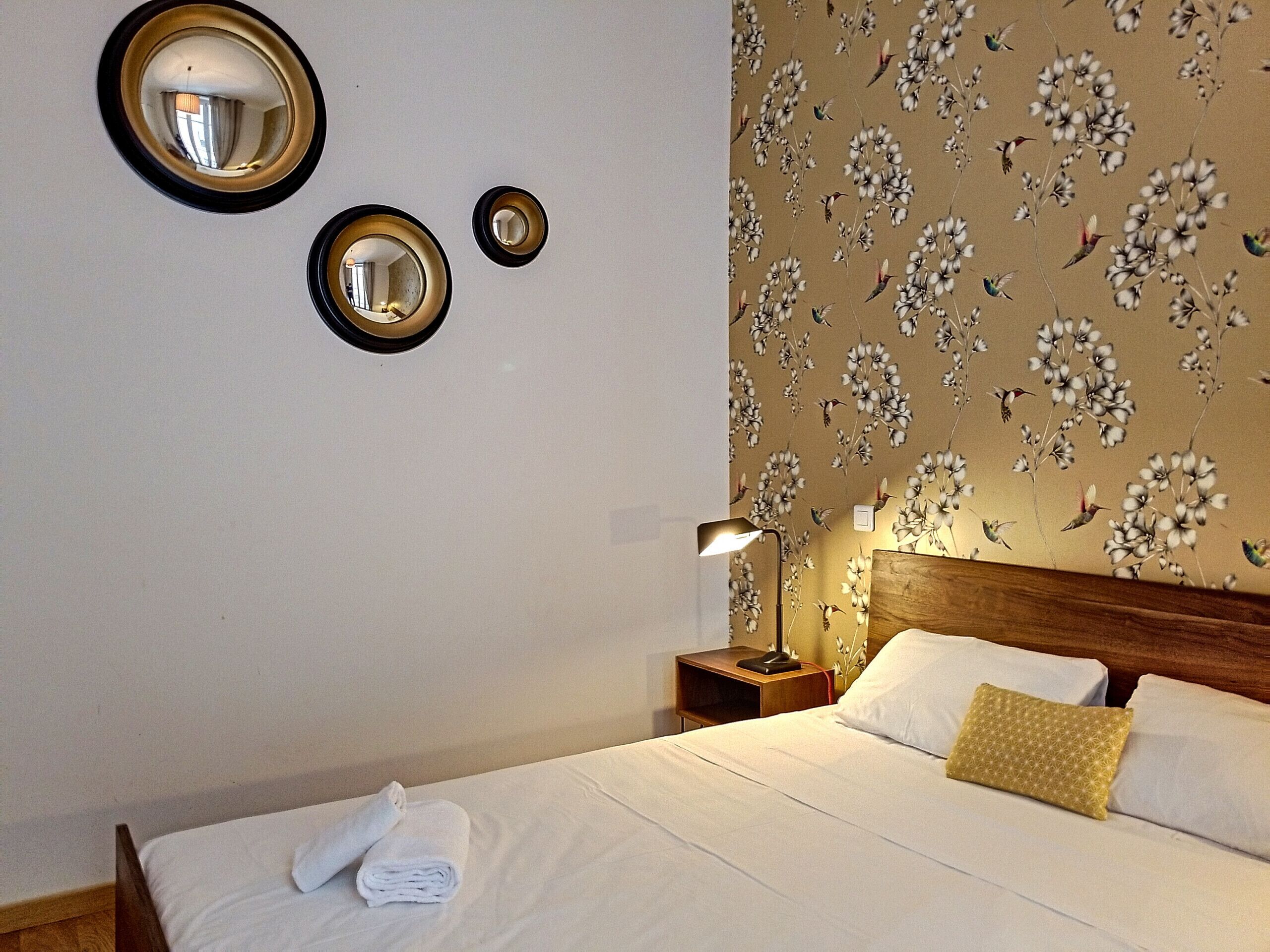 Un lit bien fait, avec du linge de lit blanc, une tête de lit en bois et un papier peint à motifs floraux. Deux miroirs sont fixés au mur, et une lampe de chevet est posée sur une table de chevet en bois.