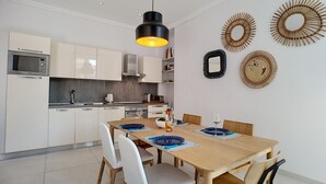 A modern kitchen with white cabinets, a built-in microwave, and a dining area with a wooden table and chairs. - N&J - COLIBRI PRESTIGE - Central - By sea (Niza)