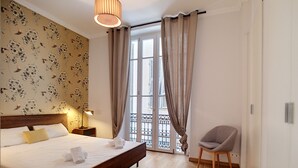 A bedroom with a wooden bed, a nightstand, a lamp, a chair, and a view of the outside through a window with curtains. - N&J - COLIBRI PRESTIGE - Central - By sea (Niza)