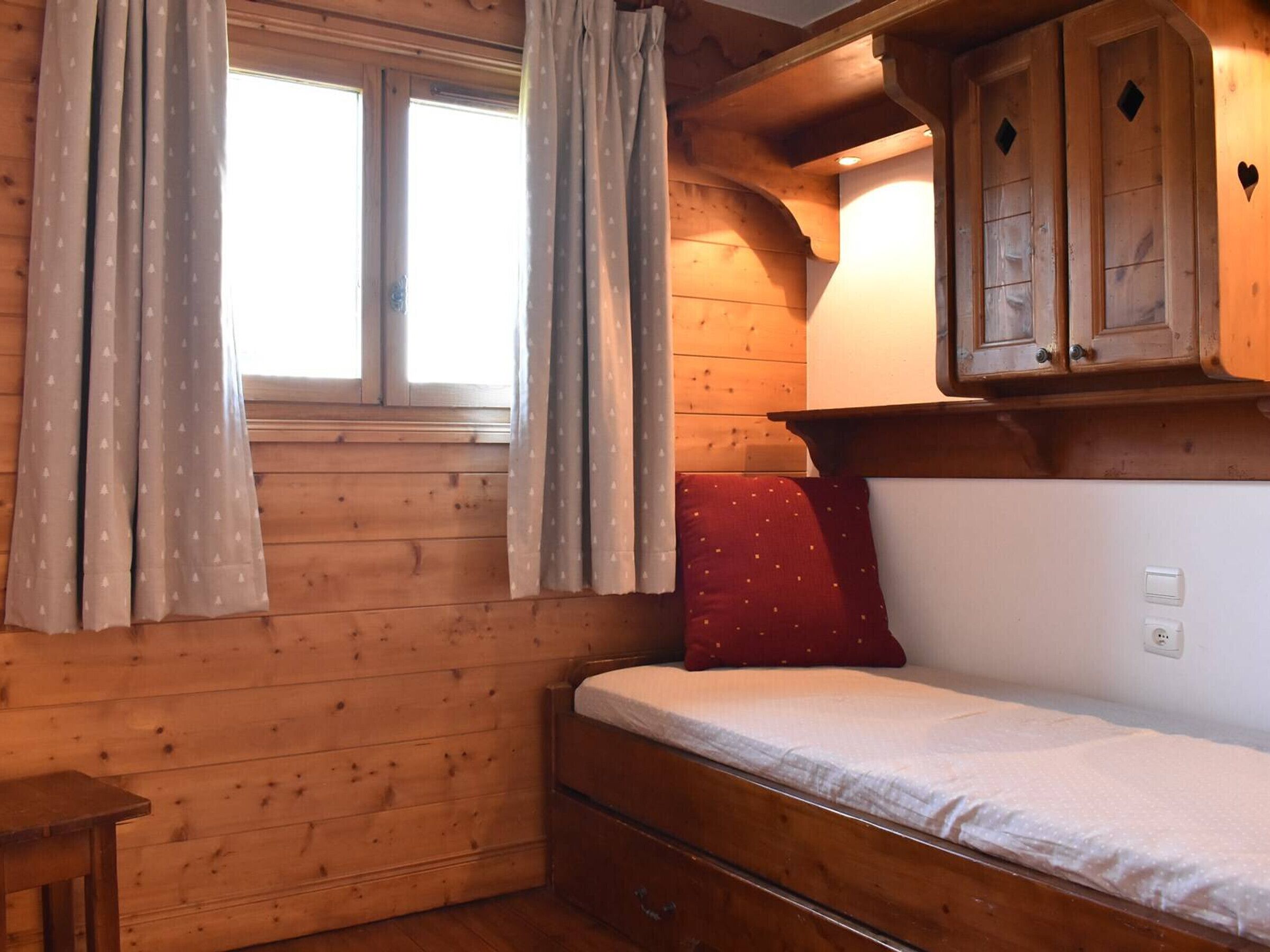A wooden cabin room with a bed, a small table, and a window with curtains.