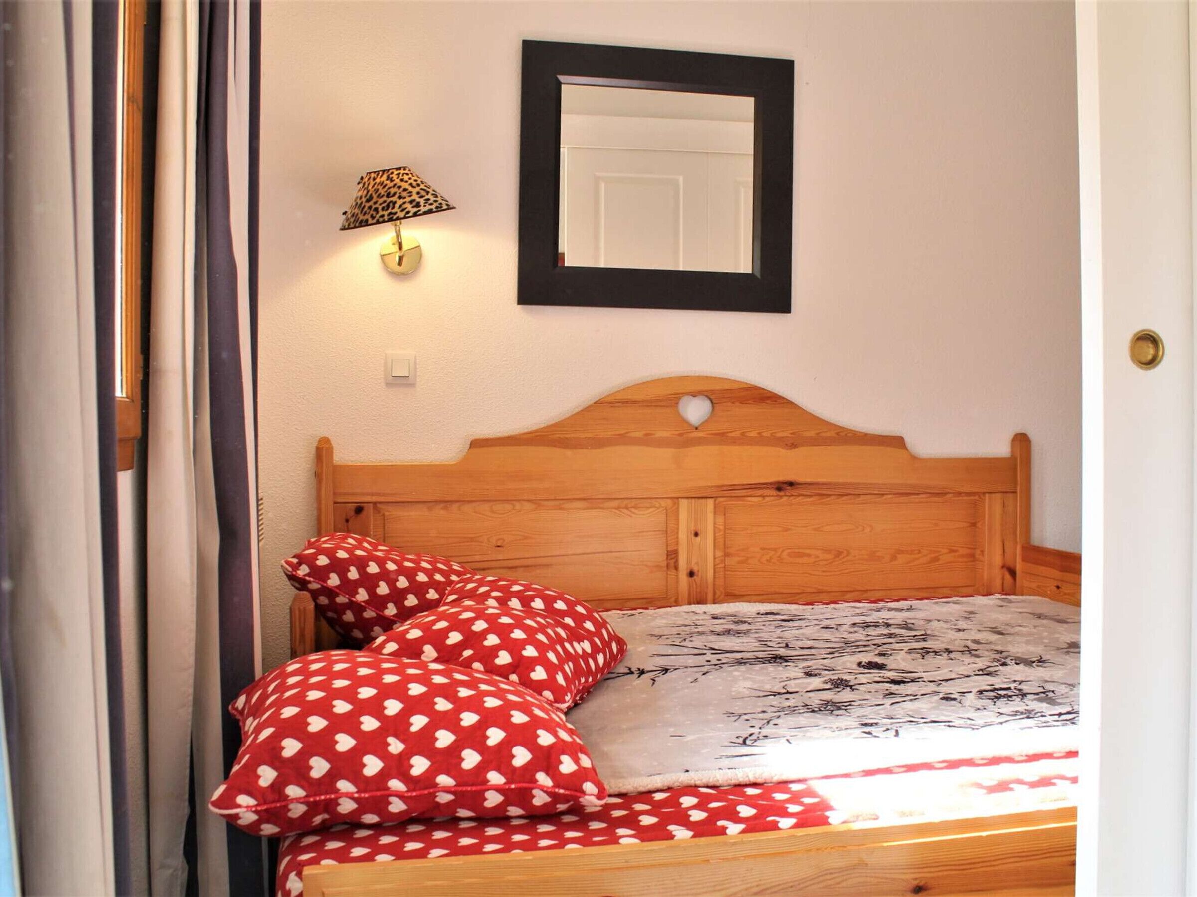A bedroom with a wooden bed, red heart-patterned pillows, a patterned bedspread, a wall-mounted lamp, and a framed mirror.