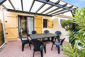 Eine Terrasse im Freien mit Tisch und Stühlen, ein überdachter Bereich mit Dach und eine Tür, die in einen Innenraum führt.