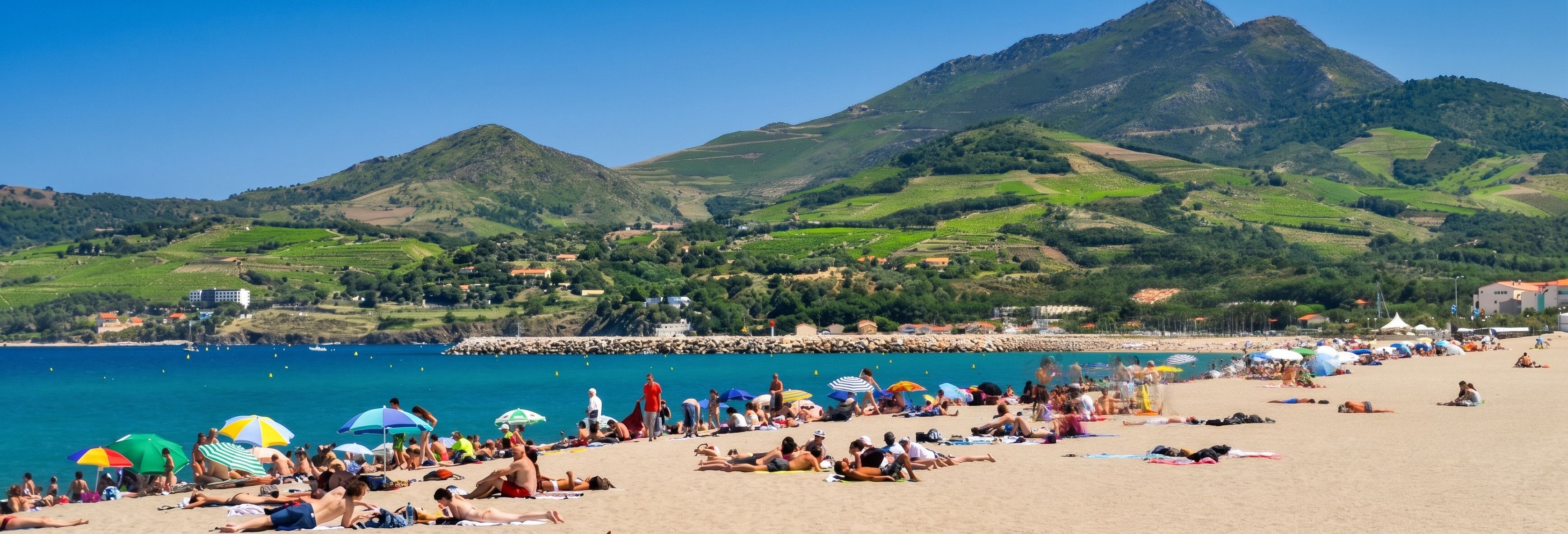 A beach with people sunbathing and swimming, a rocky breakwater, and green hills in the background.
