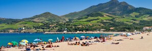 Ein Strand mit badenden Personen, einem steinernen Wellenbrecher und grünen Hügeln im Hintergrund.