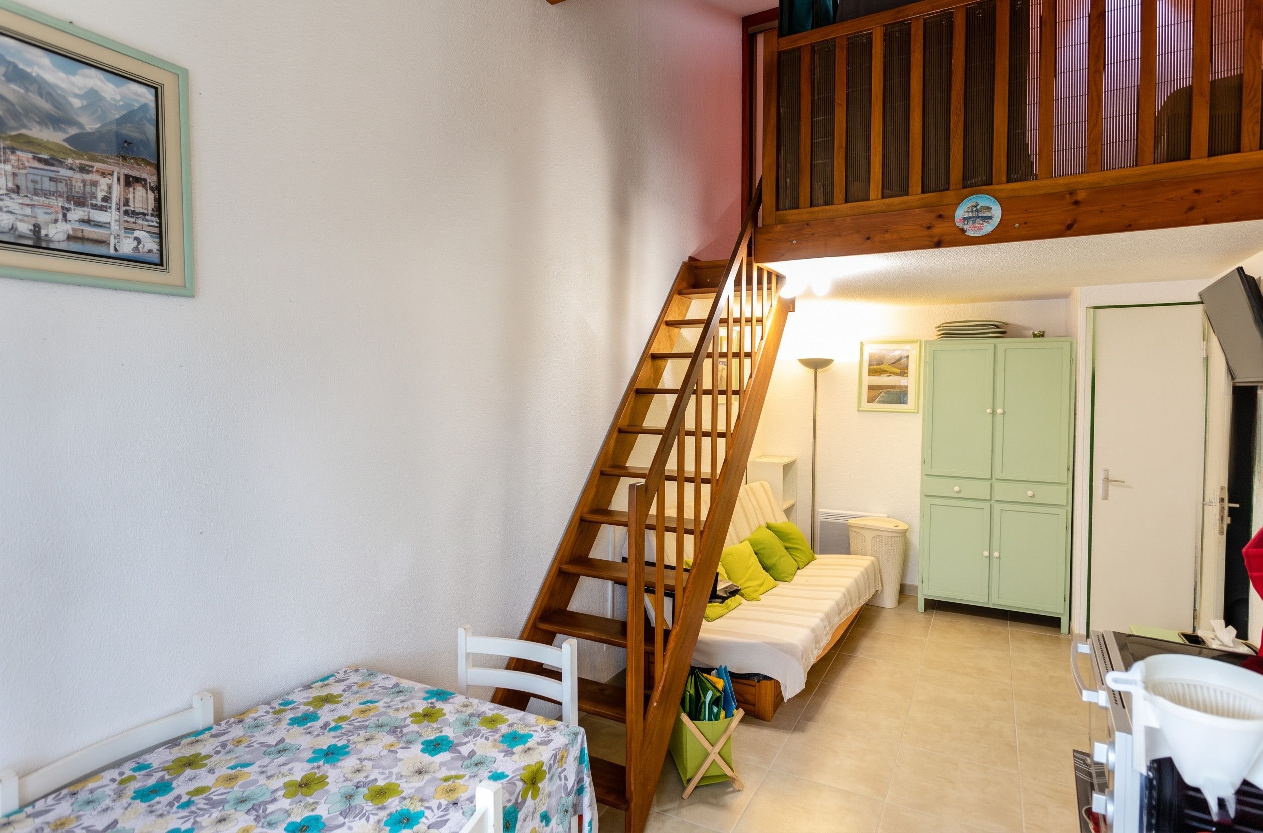 A bedroom with a wooden staircase, a bed with green pillows, a nightstand, a table with a floral tablecloth, and a green storage box.