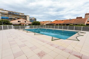 Une piscine avec une terrasse carrelée, entourée d’une clôture blanche, et des bâtiments en arrière-plan.
