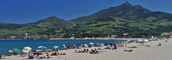Une plage où des gens se prélassent au soleil et se baignent, entourée de collines verdoyantes et sous un ciel bleu azur.