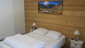 Un dormitorio con pared de madera, una cama con ropa blanca, dos mesitas de noche con lámparas y un cuadro de un paisaje montañoso.