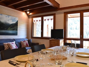 A cozy room with a dining table set for four, a sofa with patterned cushions, a television, and large windows with curtains.