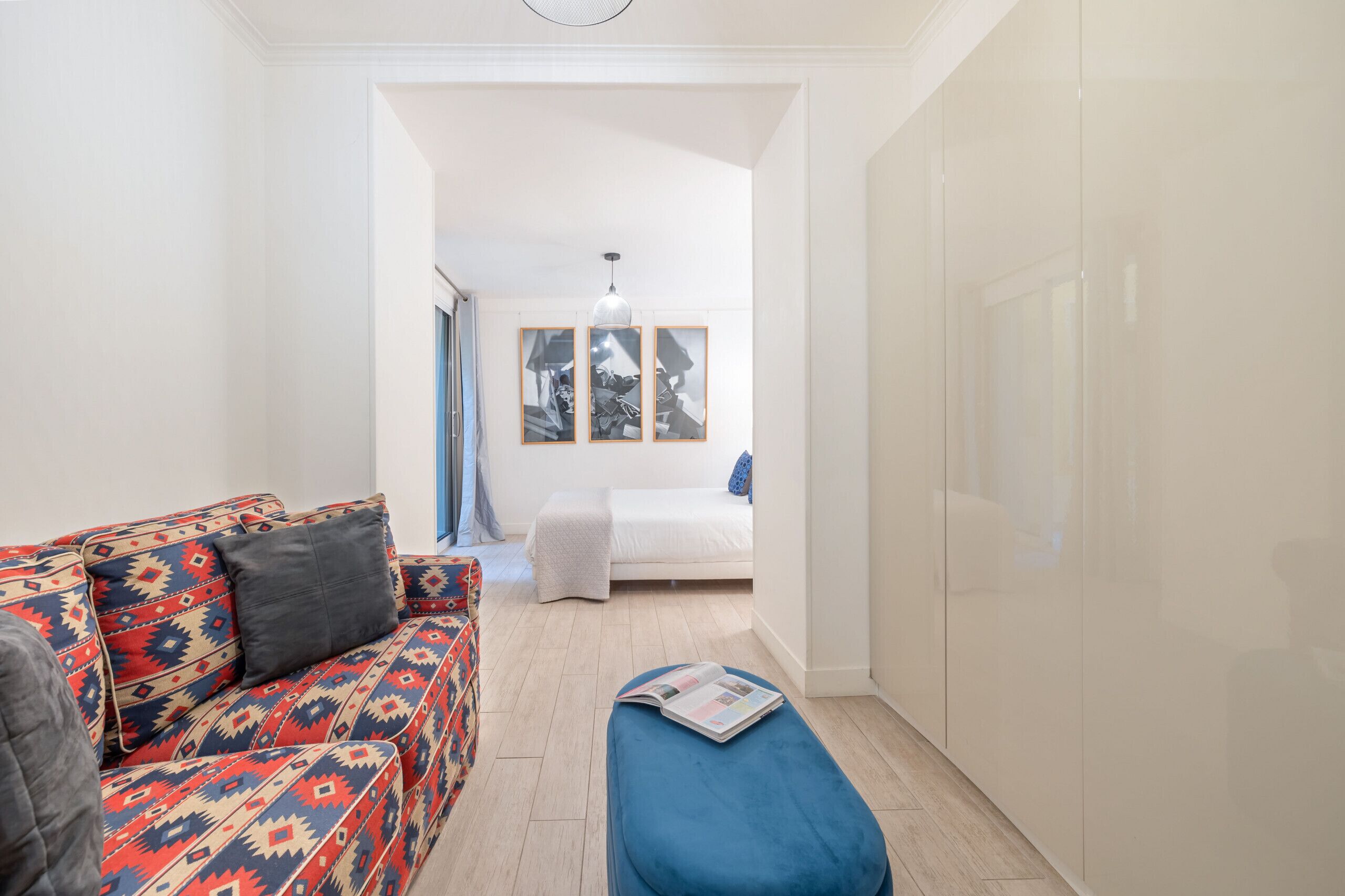 A narrow hallway with a patterned sofa, a blue ottoman, and a bed with a white mattress and blue bedding.