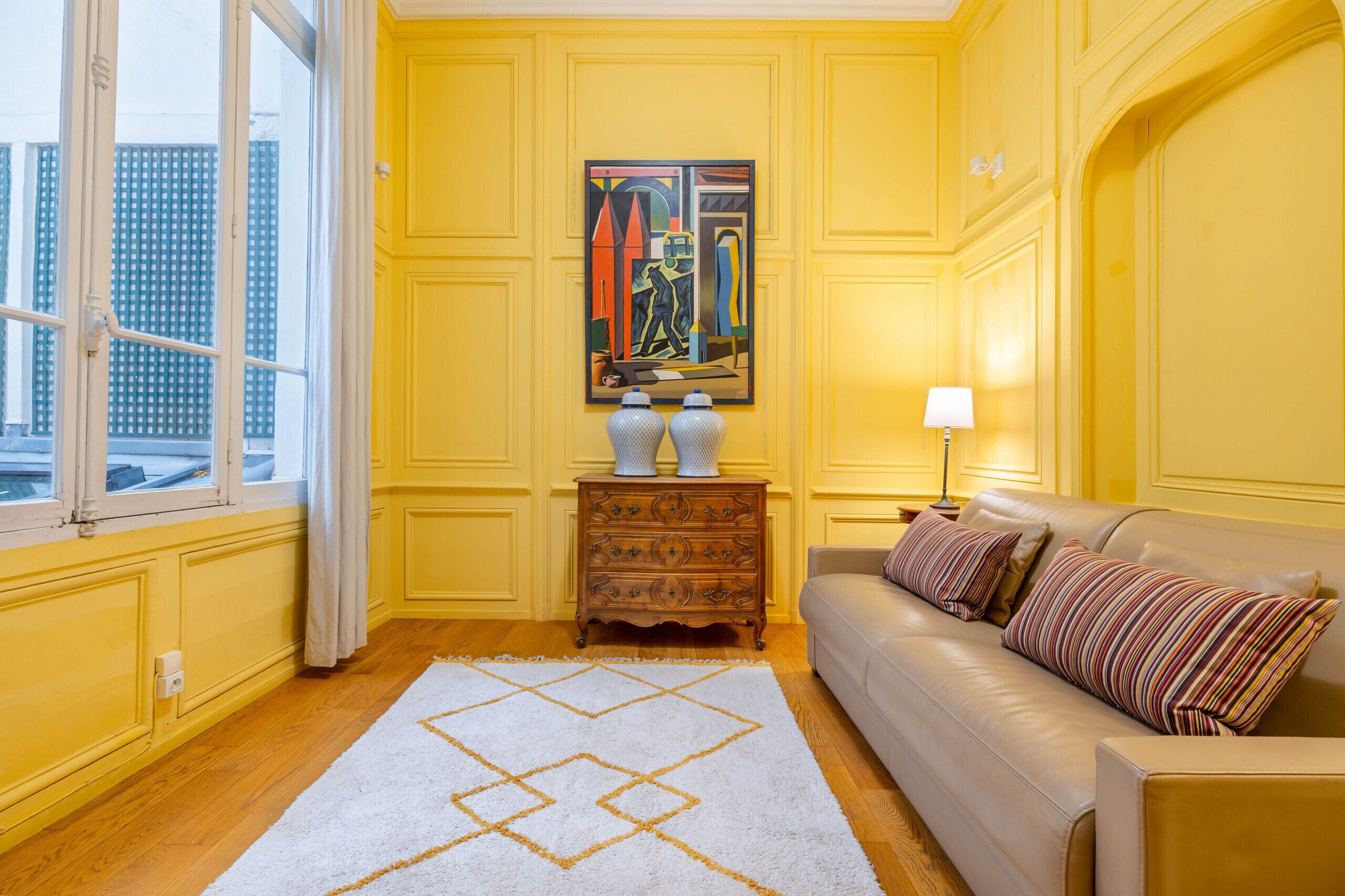 A room with yellow walls, a beige sofa, a patterned rug, a painting, and a window with curtains.