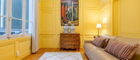 A room with yellow walls, a beige sofa, a patterned rug, a painting, and a window with curtains.