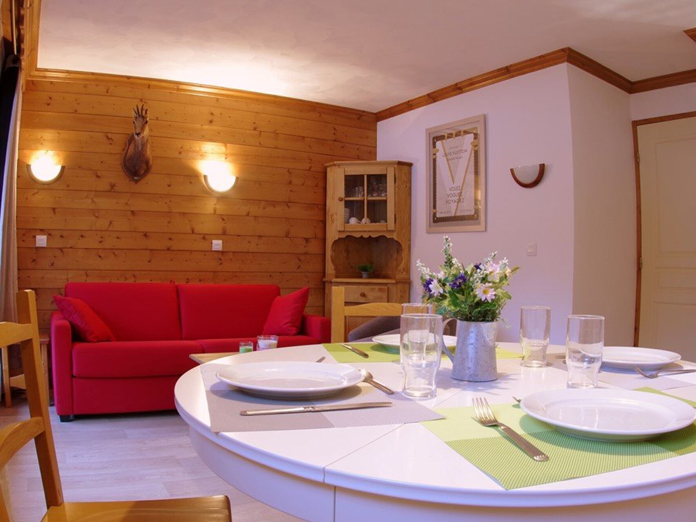 A cozy room with a wooden wall, a red sofa, a dining table set with plates, glasses, and cutlery.