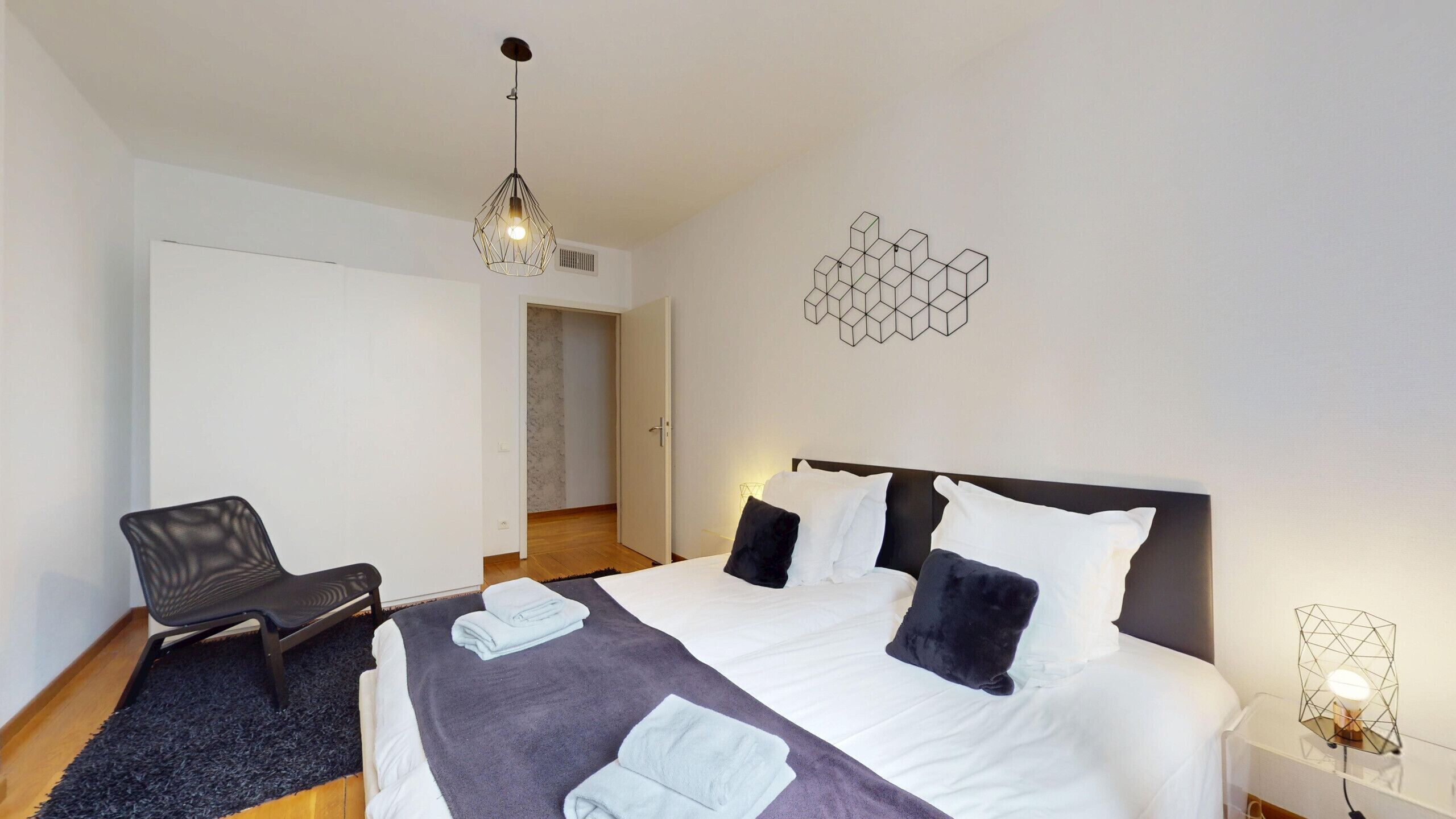 A modern hotel room with a large bed, a chair, a desk, and a geometric wall decoration.