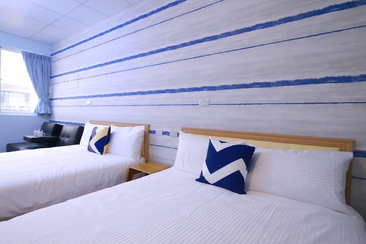 A hotel room with two beds, each with a wooden headboard, white bedding, and blue and white pillows. There is a small wooden nightstand between the beds, a black leather chair, and a window with a curtain.