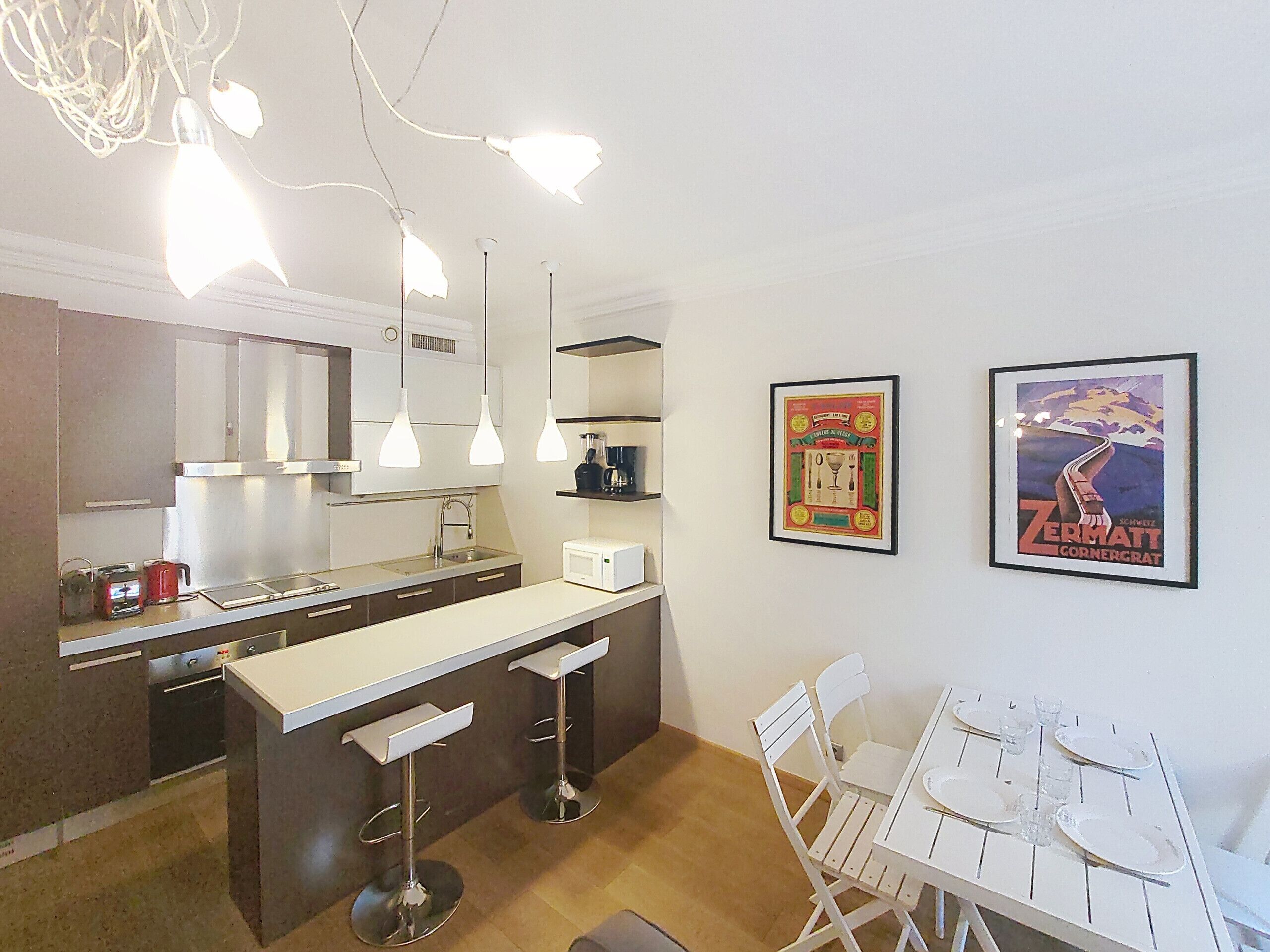 A modern kitchen with a central island, bar stools, a dining table with chairs, pendant lights, and two framed pictures on the wall.
