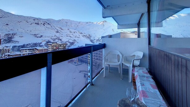 Een balkon met uitzicht op met sneeuw bedekte bergen, een houten tafel met een geruit tafelkleed en twee witte plastic stoelen.