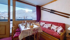 Een knus vertrek met een houten eettafel, een rode bank met opvallende kussens, en een uitzicht op besneeuwde bergen door grote ramen.