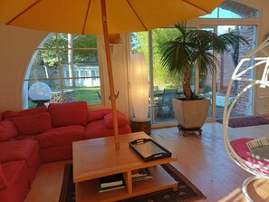 A cozy living room with a red sofa, a wooden coffee table, a hanging chair, and a large window with a view of greenery outside.