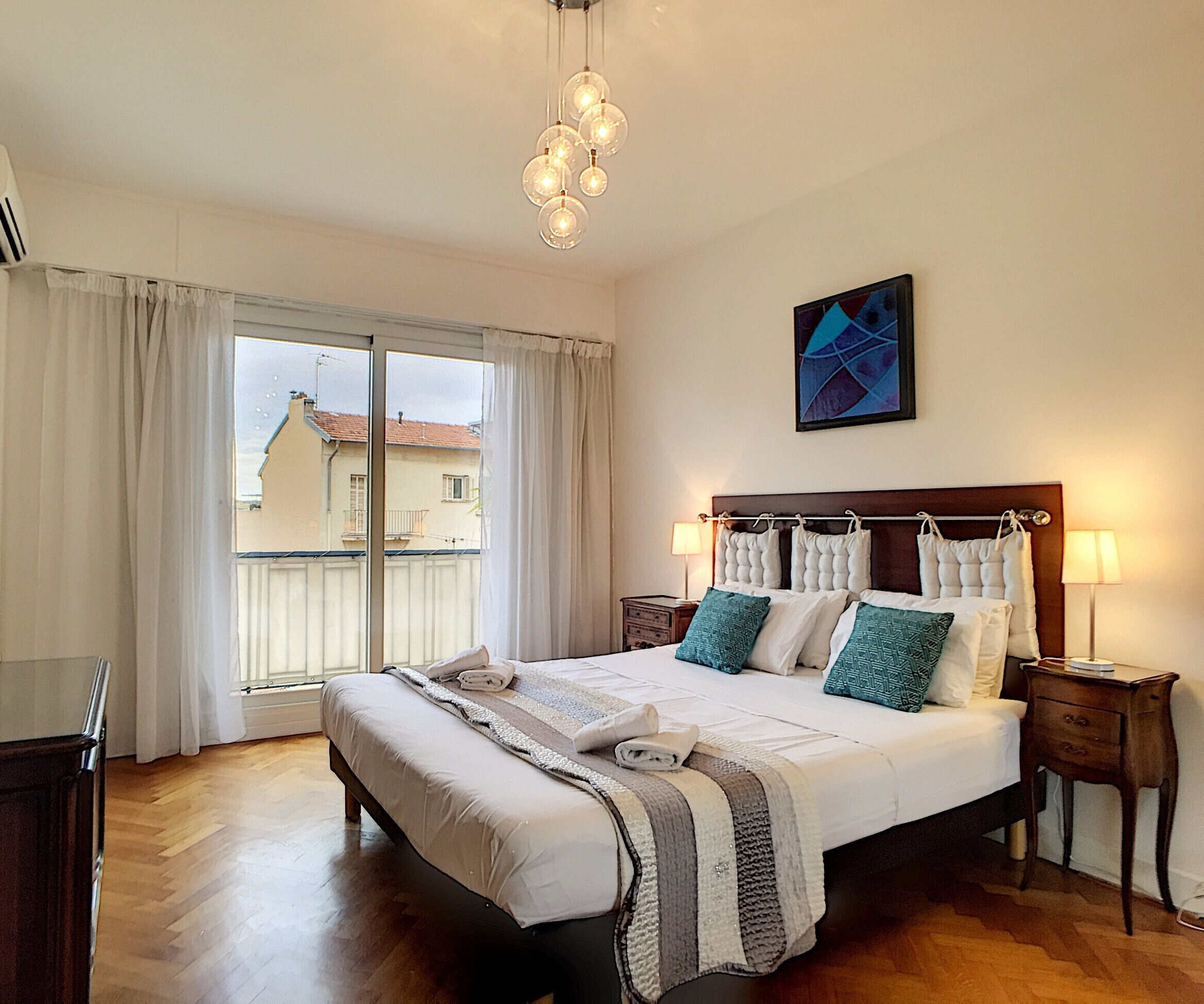 A hotel room with a large bed, a wooden floor, a window with curtains, and a wall-mounted artwork.
