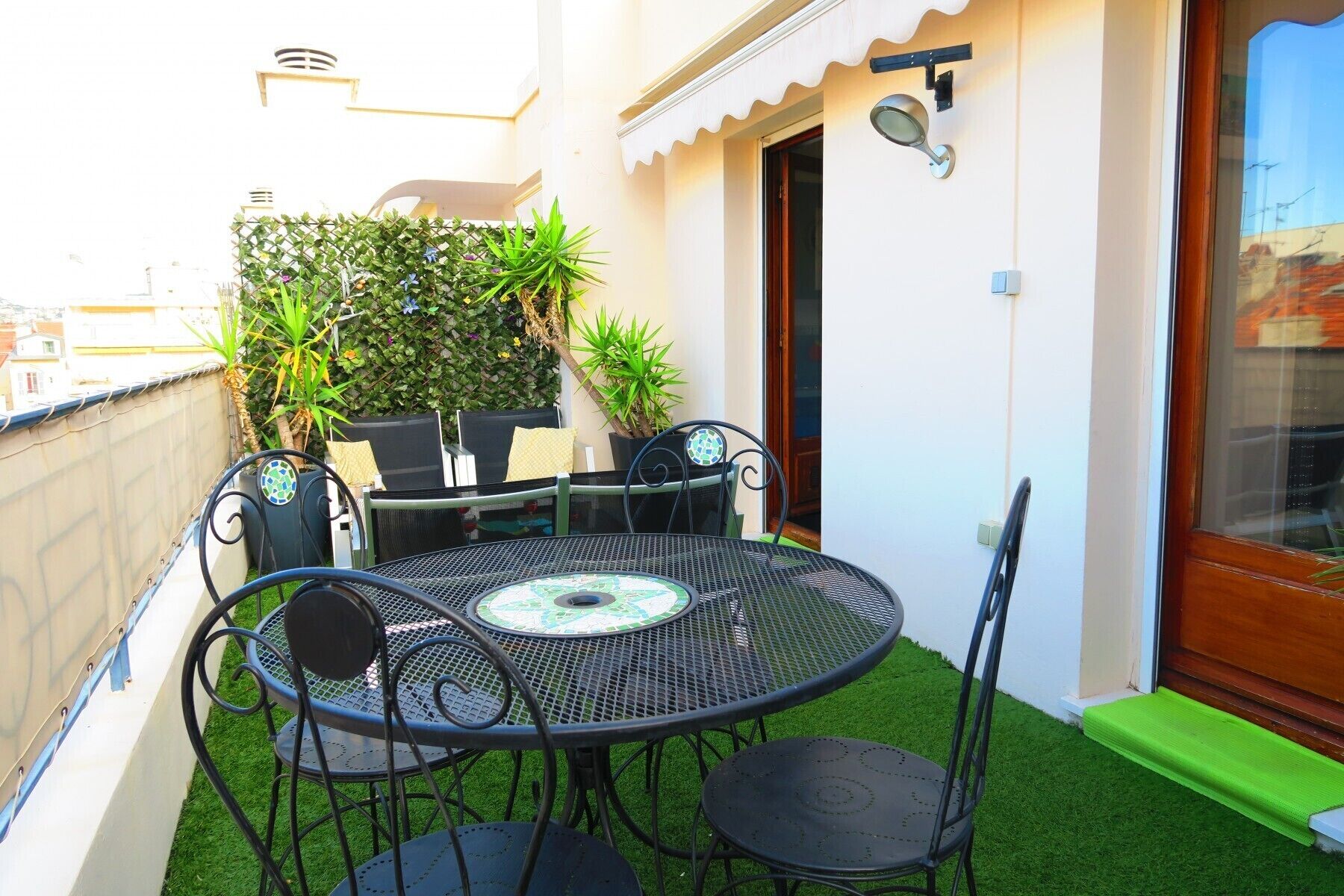 A small outdoor patio with a round table and four chairs, surrounded by potted plants and a white wall.