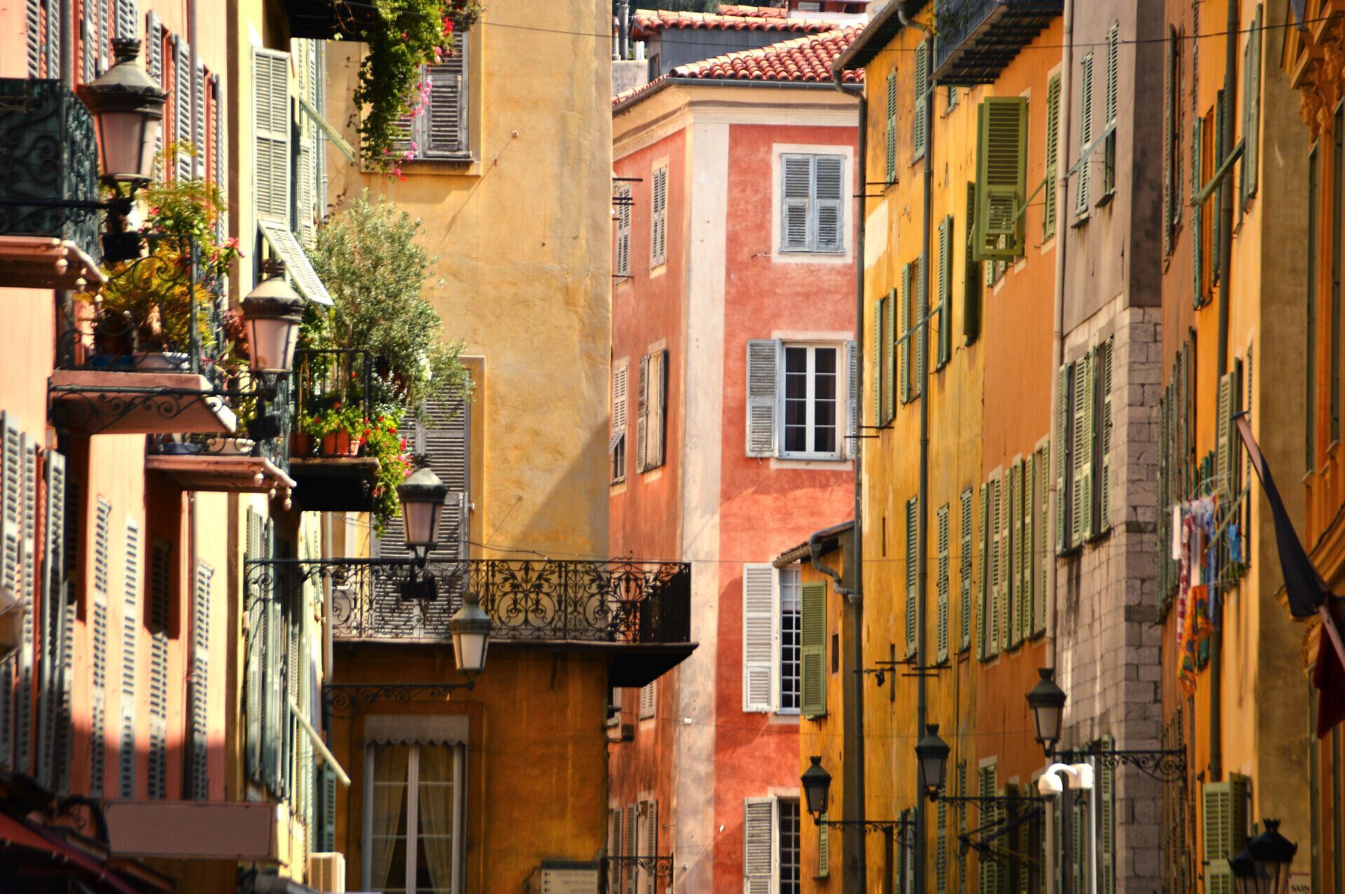 A narrow street with colorful buildings, balconies with plants, and traditional street lamps.