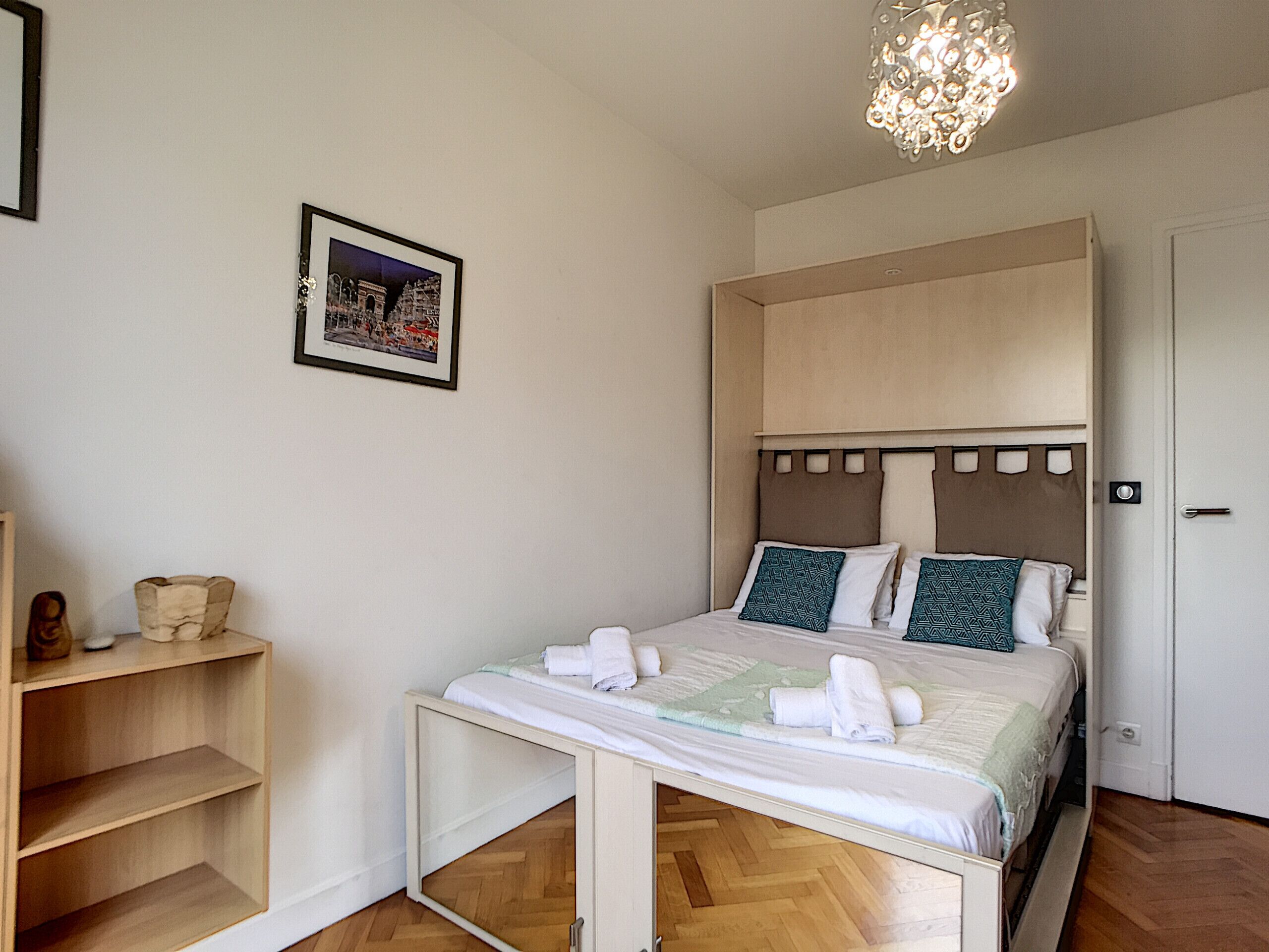 A bedroom with a bed, a bedside table, a shelf, a framed picture, and a door.