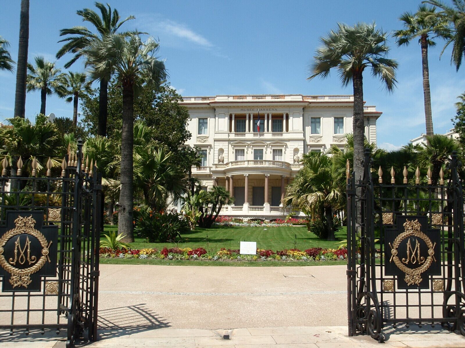 A grand building with a formal entrance, palm trees, and a well-maintained garden.