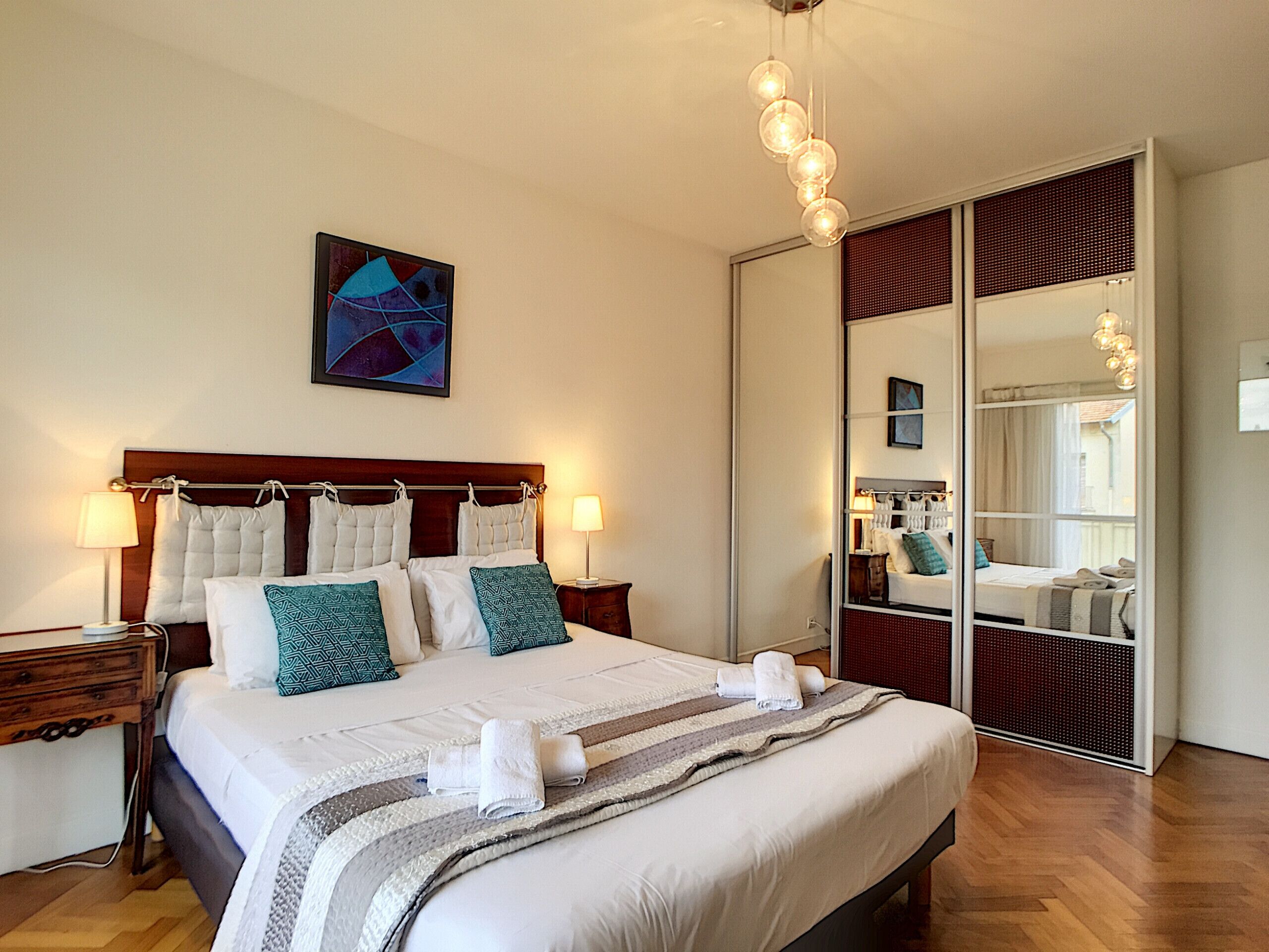 Una habitación de hotel ordenada con una cama grande, lámparas de noche, un espejo y un cuadro en la pared.