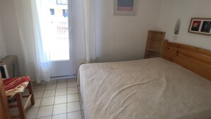 A bedroom with a bed, a chair, a radiator, a window with curtains, and a small shelf. - 3-Bedroom Villa + Mezzanine for 6 in Port Leucate with Parking (Port Leucate)