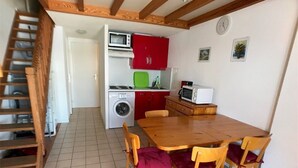 A compact kitchen with a washing machine, a microwave, and red cabinets. There is a wooden dining table with four chairs, and a staircase is visible to the left. - 3-Bedroom Villa + Mezzanine for 6 in Port Leucate with Parking (Port Leucate)
