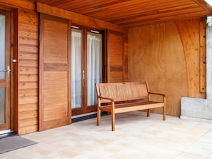 Una panchina di legno in un'area esterna coperta, con pavimento in piastrelle, pareti di legno e una porta scorrevole in vetro.