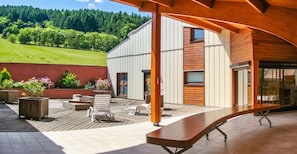 A modern outdoor patio with a wooden deck, seating area, and a view of a forested hillside.