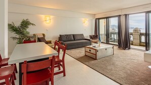 Un soggiorno moderno con zona pranzo, un divano grigio, sedie rosse, un tavolo bianco e un'ampia finestra che si affaccia su uno specchio d'acqua e edifici.