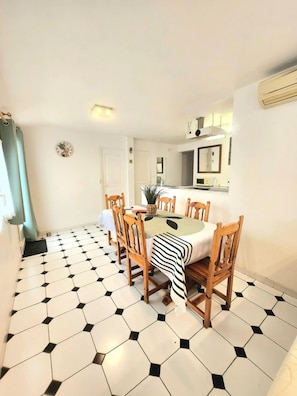 Uma área de refeições com piso de azulejos hexagonais, cadeiras de madeira, uma mesa com toalha listrada e uma cozinha com fogão e pia.