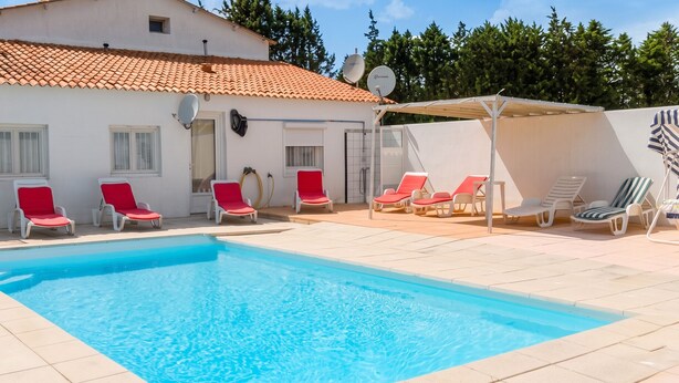A swimming pool with red and white lounge chairs, a covered area, and a white building with a tiled roof. - Homerez - Villa in Meynes (Meynes)
