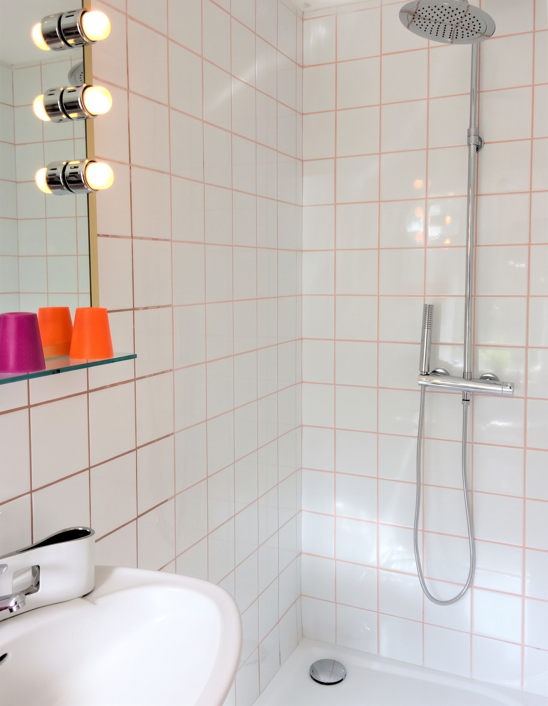 Een badkamer met een wit wastafel, een spiegel met verlichting, een douche met handdouche, en een glazen plank met oranje en paarse bekers.
