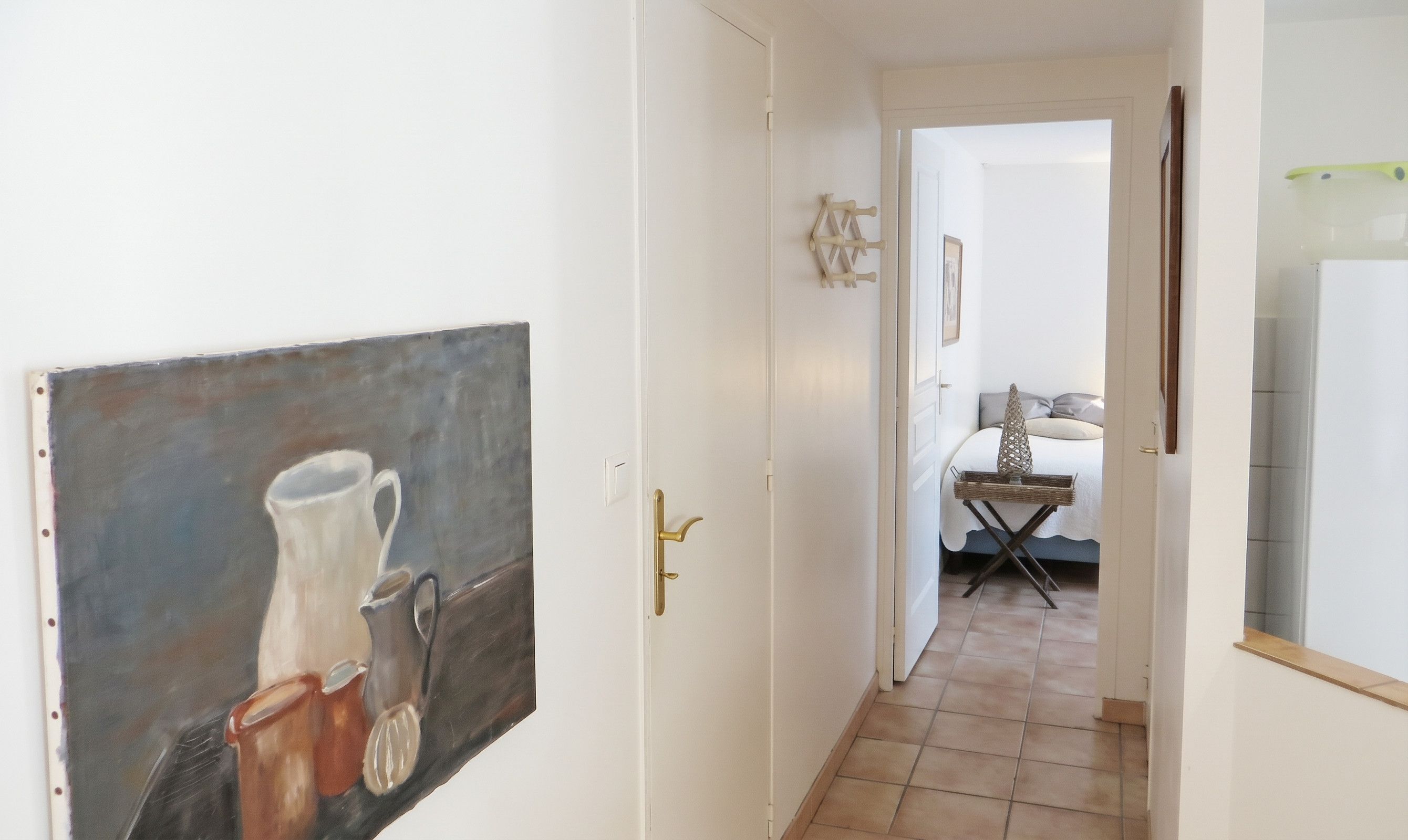 Un couloir orné d’un tableau représentant des vases, un lit recouvert d’une couvre-lit à motifs et une petite table sur laquelle se trouve un panier.