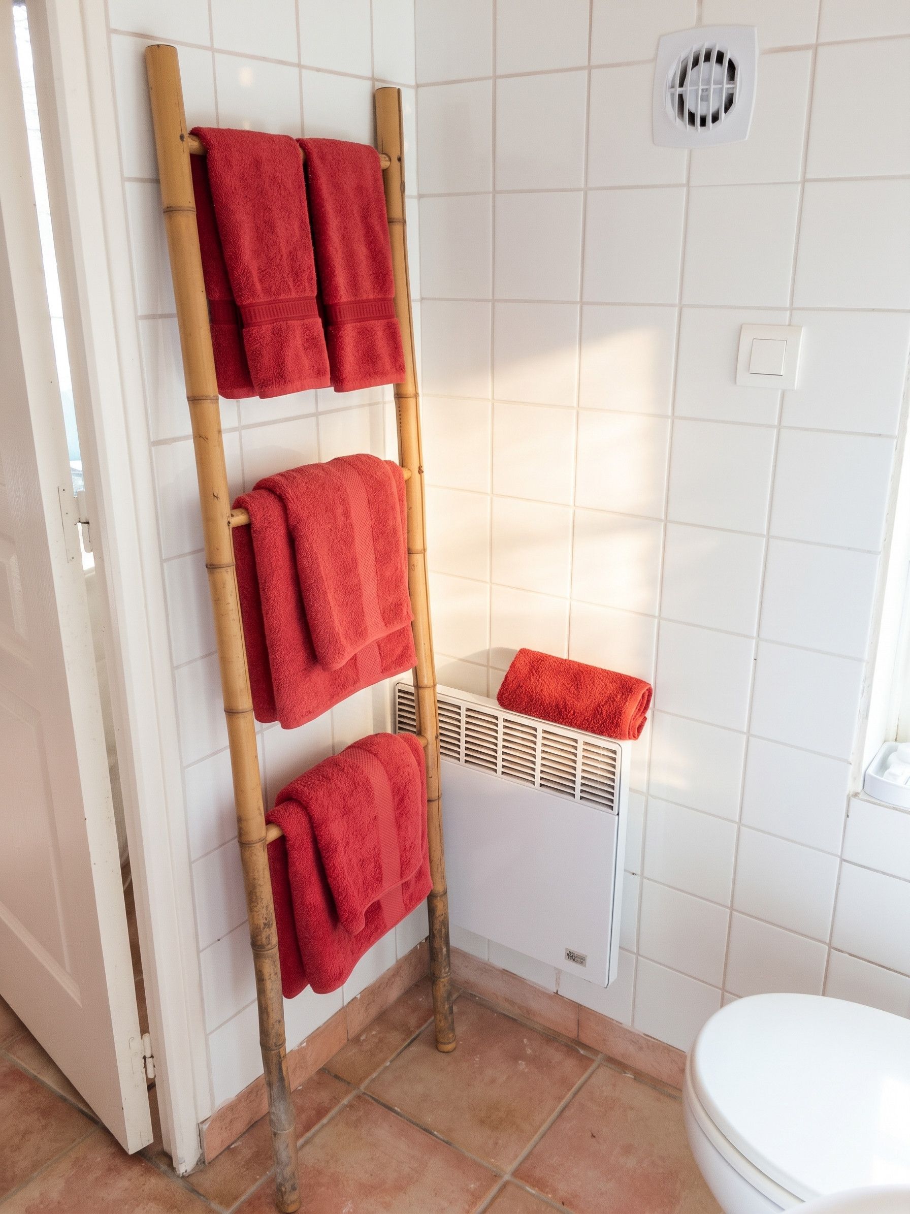 Une salle de bain avec un porte-serviettes en bambou sur lequel sont disposées trois serviettes, des toilettes blanches et une grille d’aération au mur.