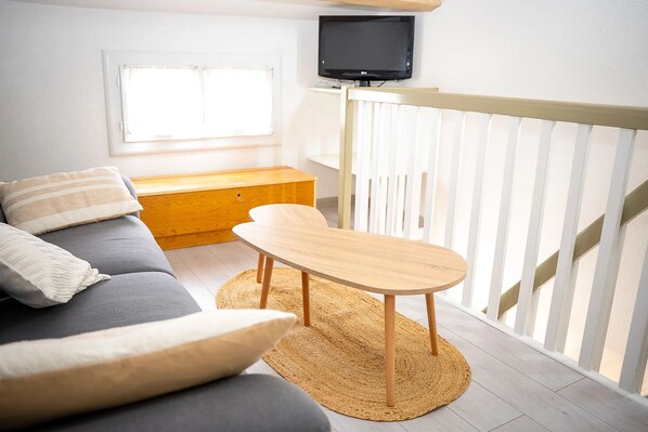 A room with a sofa, a small wooden table, a TV mounted on the wall, and a window with blinds.
