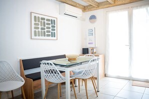 A dining area with a glass table, white chairs, a wooden bench, a framed abstract artwork, an air conditioning unit, a clock, and a door with curtains.